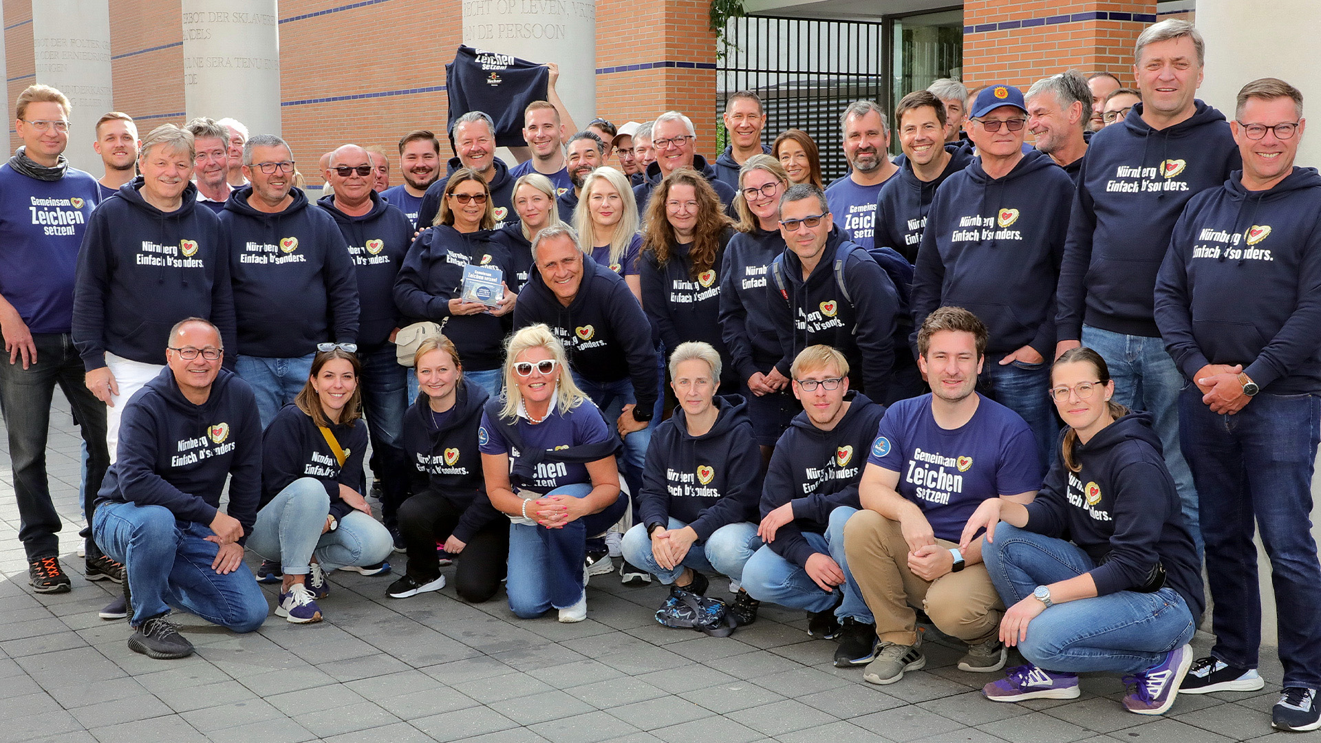 Eine Gruppe von Personen in blauen Kapuzenpullis steht lächelnd vor einem Gebäude.