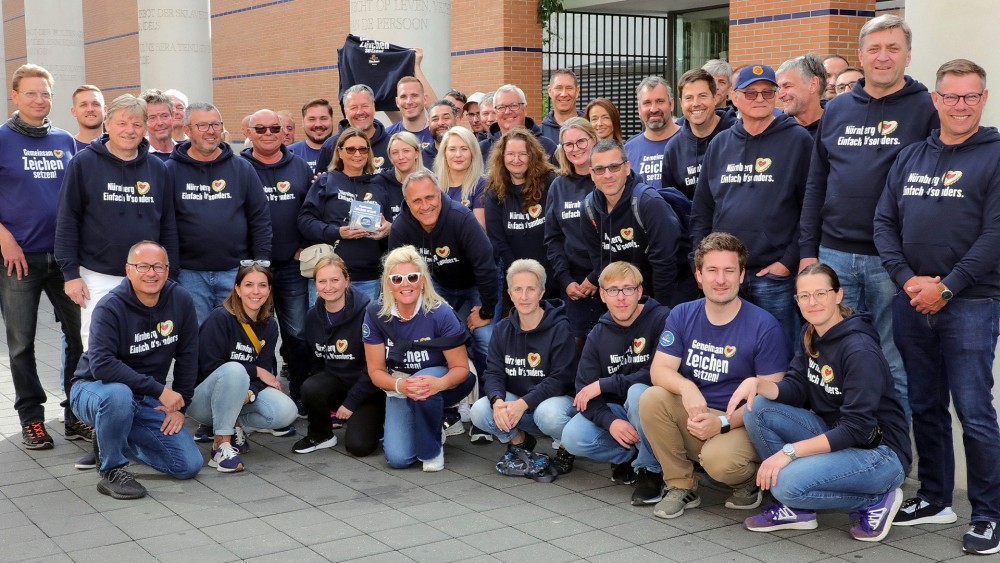 Eine Gruppe von Personen in blauen Kapuzenpullis steht lächelnd vor einem Gebäude.