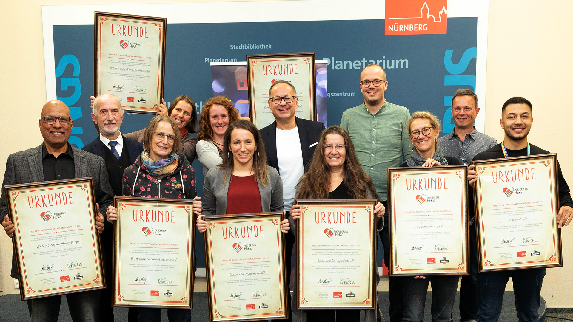 Elf Personen stehen nebeneinander und halten stolz ihre Urkunden, sie lächeln in die Kamera.