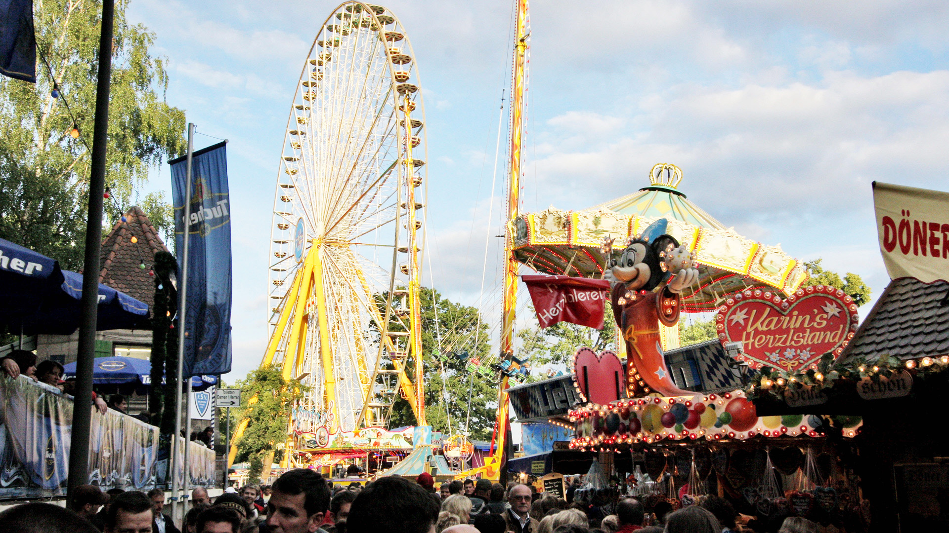 Ein belebter Jahrmarkt mit einem großen Riesenrad und verschiedenen Ständen bei Tageslicht.