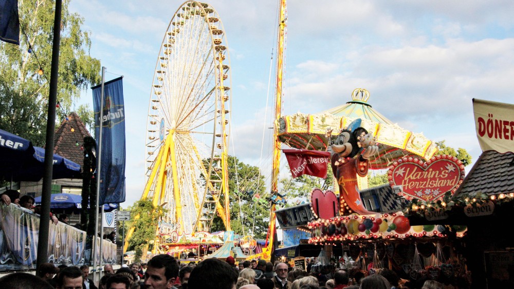 Ein belebter Jahrmarkt mit einem großen Riesenrad und verschiedenen Ständen bei Tageslicht.