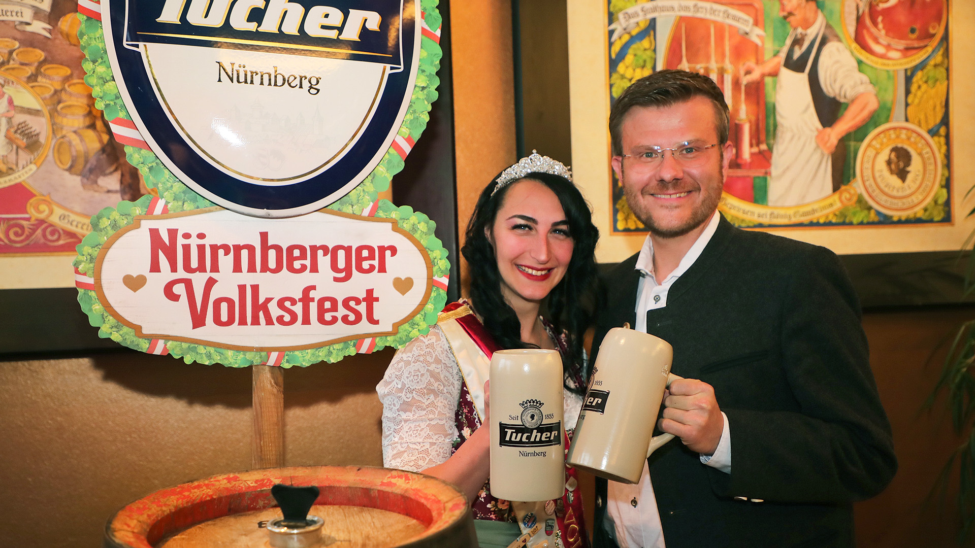 Zwei Personen lächeln und halten große Bierkrüge, umgeben von Volksfestdekorationen.