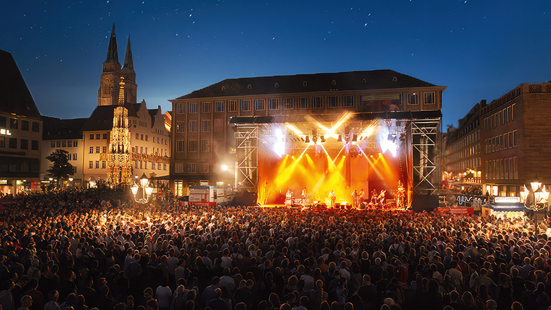 Eine große Menschenmenge steht bei Nacht auf einem beleuchteten Platz vor einer Bühne mit Live-Musik.
