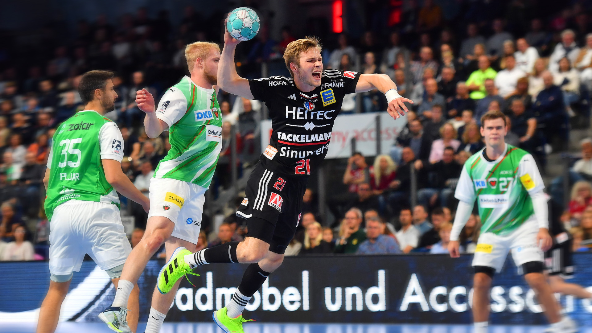 Drei Handballspieler kämpfen um den Ball bei einem Spiel in einer großen Halle. Der Spieler in der Mitte springt und wirft den Ball.