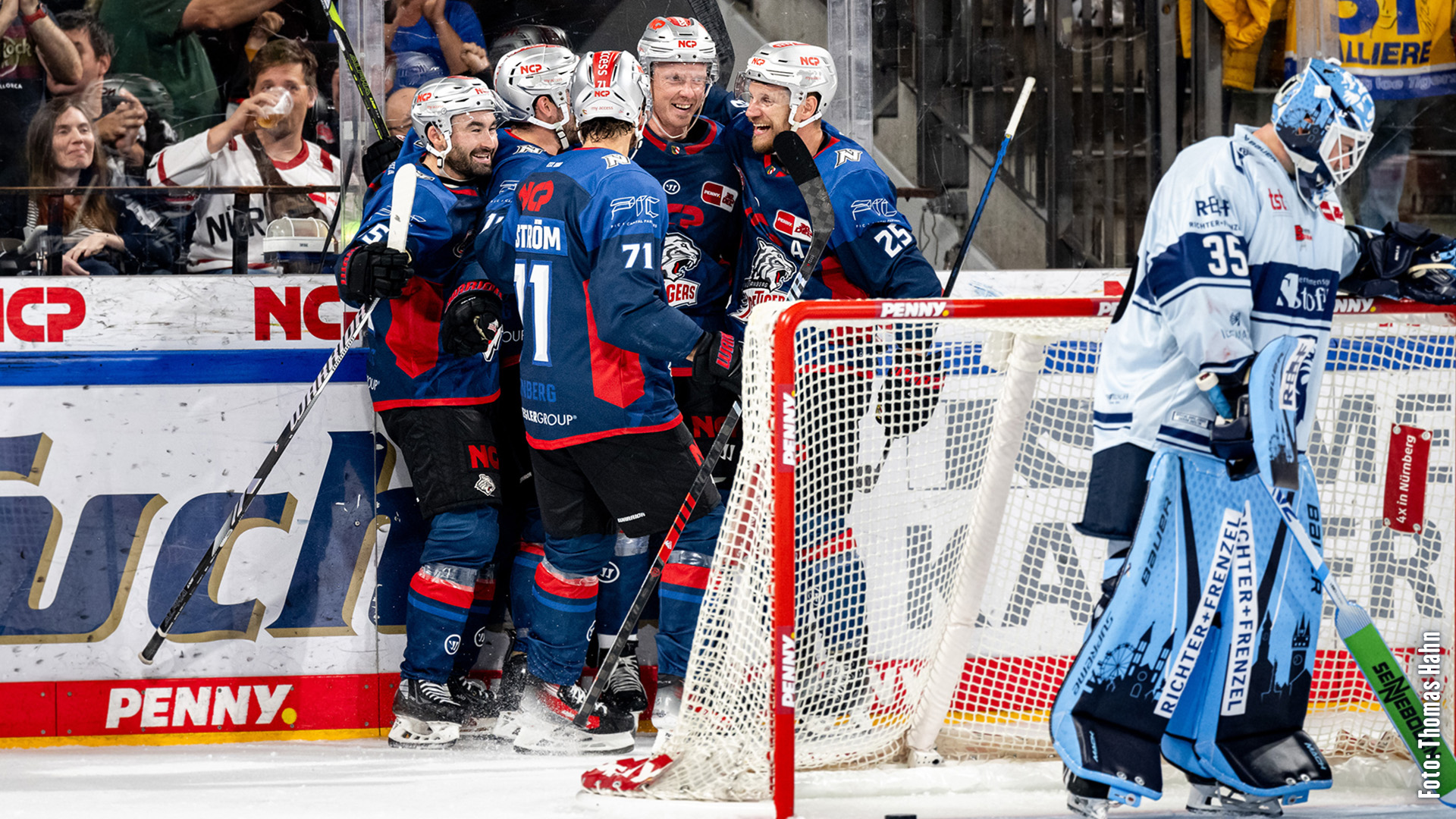 Vier Eishockeyspieler in blauen Trikots feiern ein Tor, während ein Torwart daneben steht.