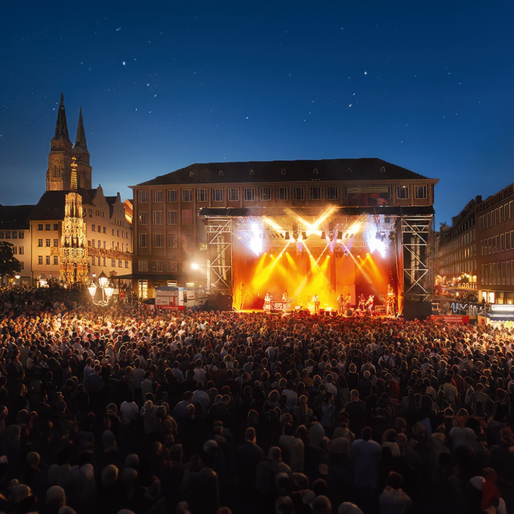 Eine große Menschenmenge steht abends auf einem Platz vor einer beleuchteten Bühne mit Musikern.